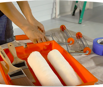 Person holding two paint rollers next to two paint brushes in a roller tray
